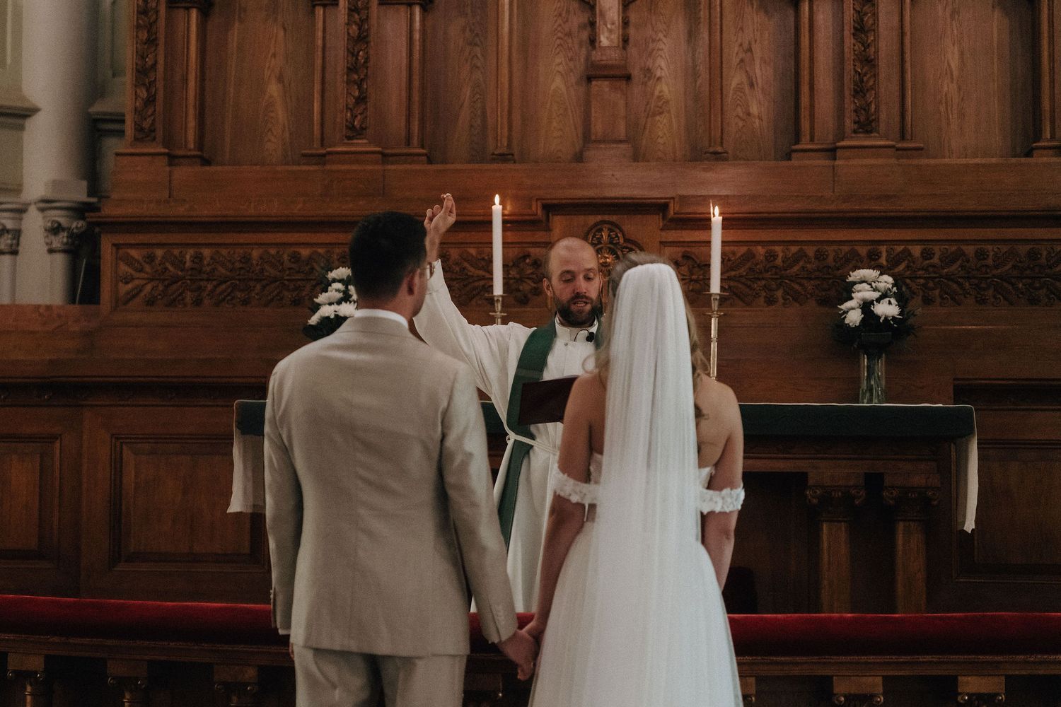 Officiant raising hand in blessing over bride and groom holding hands at wooden church altar with candles.