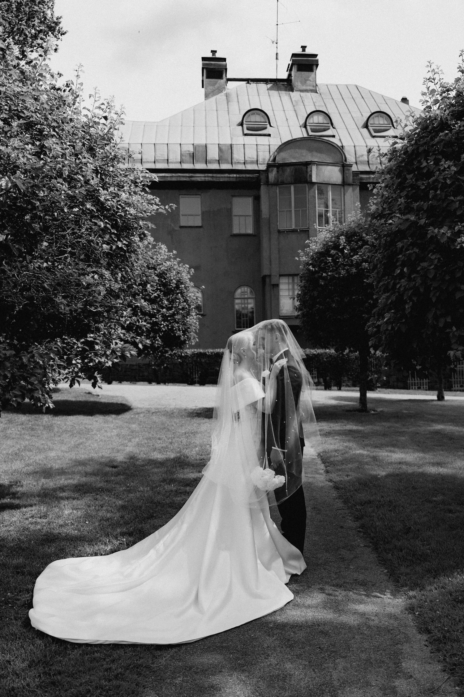 Black and white photo of bride and groom kissing under flowing veil in front of a historic manor.