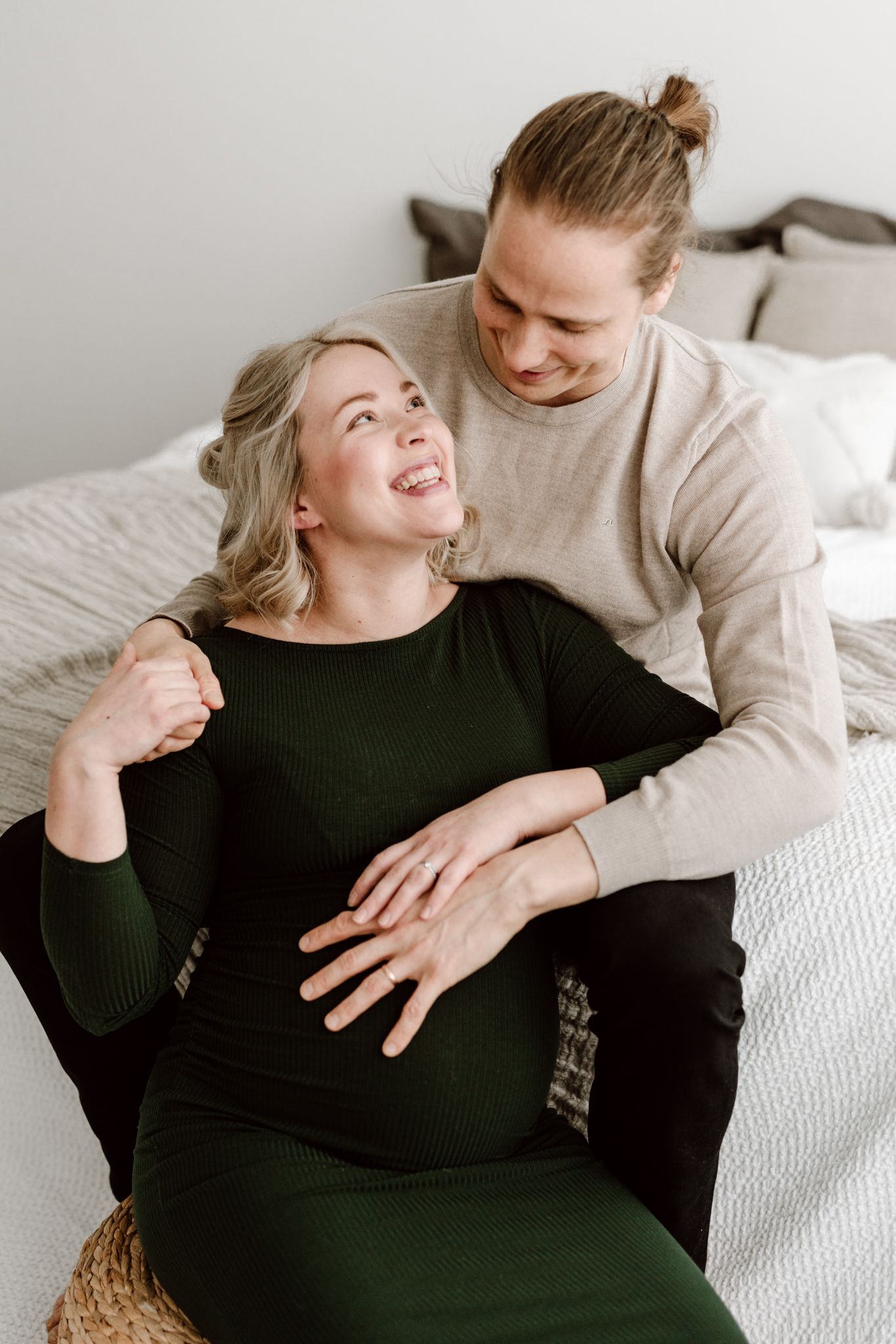 Happy pregnant couple sitting on bed, woman in green dress laughing while partner embraces her.