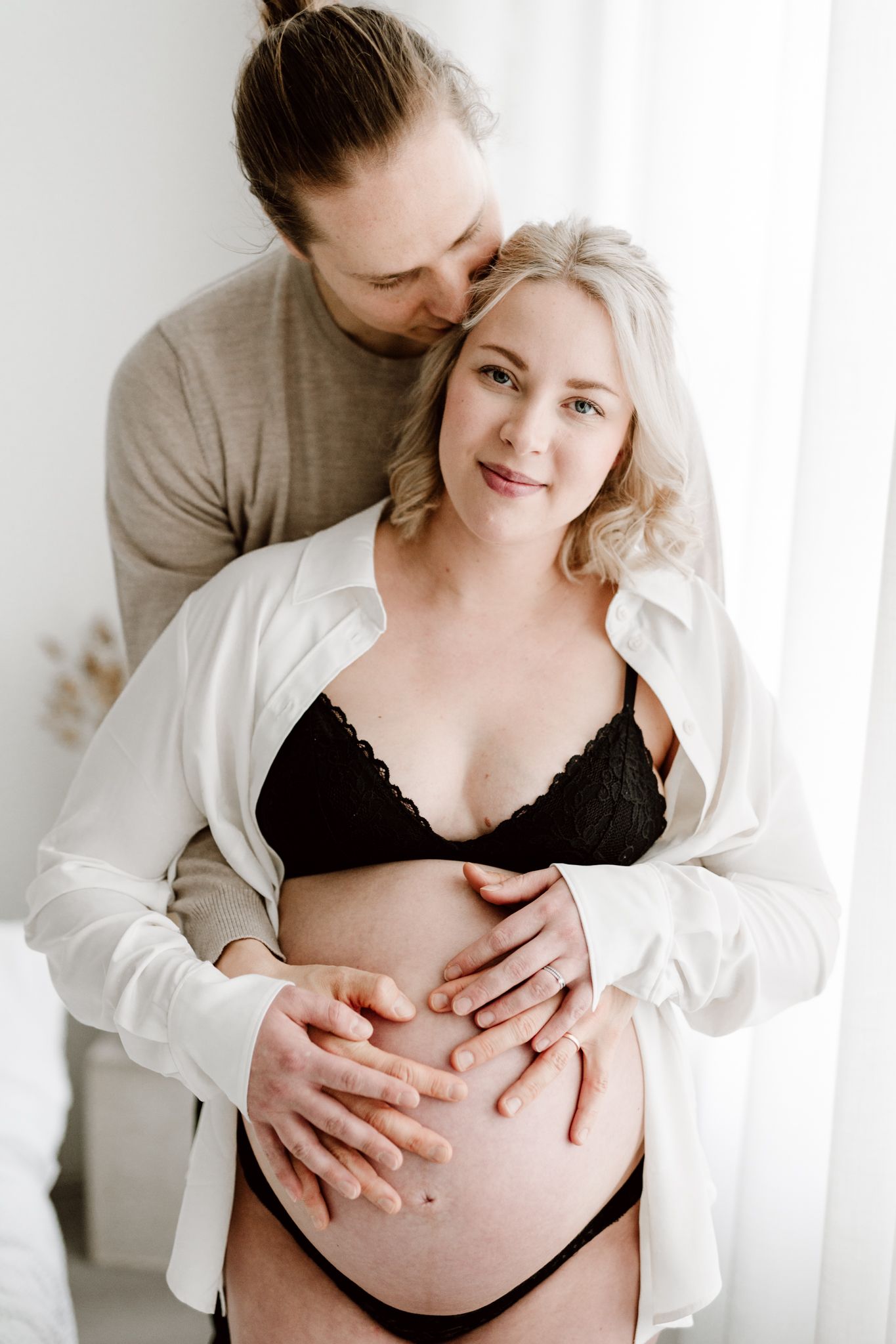 Loving man kisses pregnant blonde woman's head while embracing her baby bump in bright white room.
