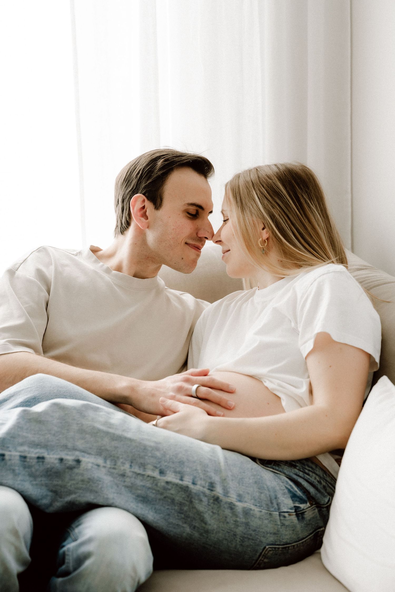 Couple in white shirts sitting on sofa touching pregnant belly nose to nose.
