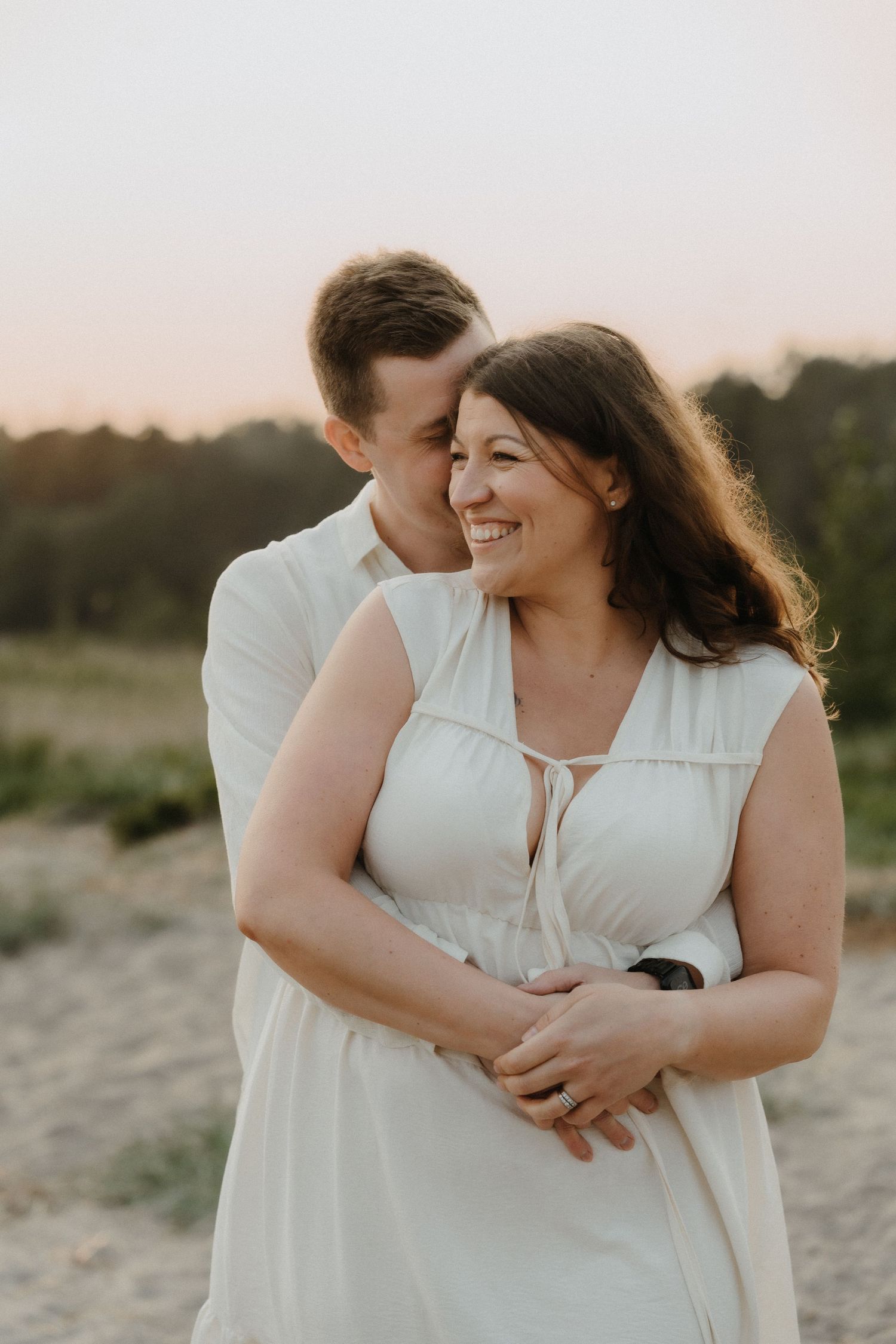 A smiling couple embraces tenderly at sunset, both wearing white in a serene outdoor setting.