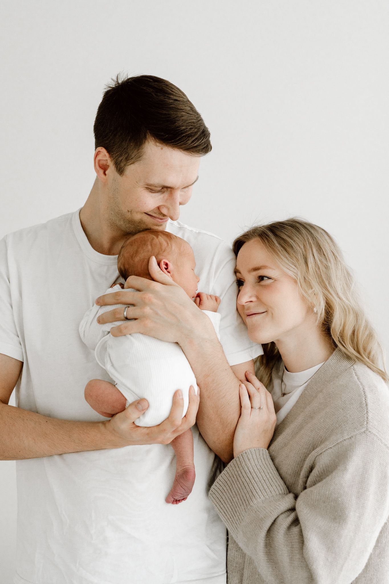 New parents in white and beige outfits lovingly gazing at their newborn baby together.