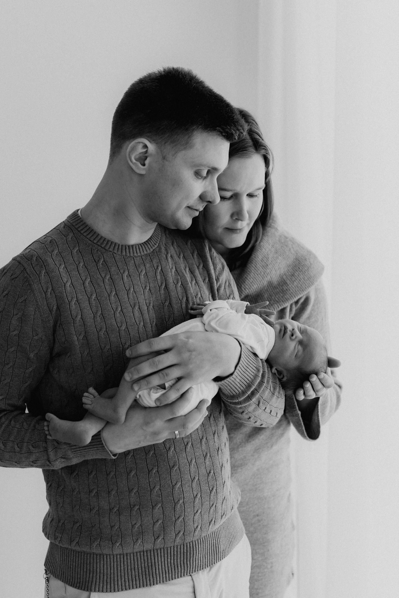 Black and white portrait of parents in knitwear tenderly holding their newborn baby by a window.