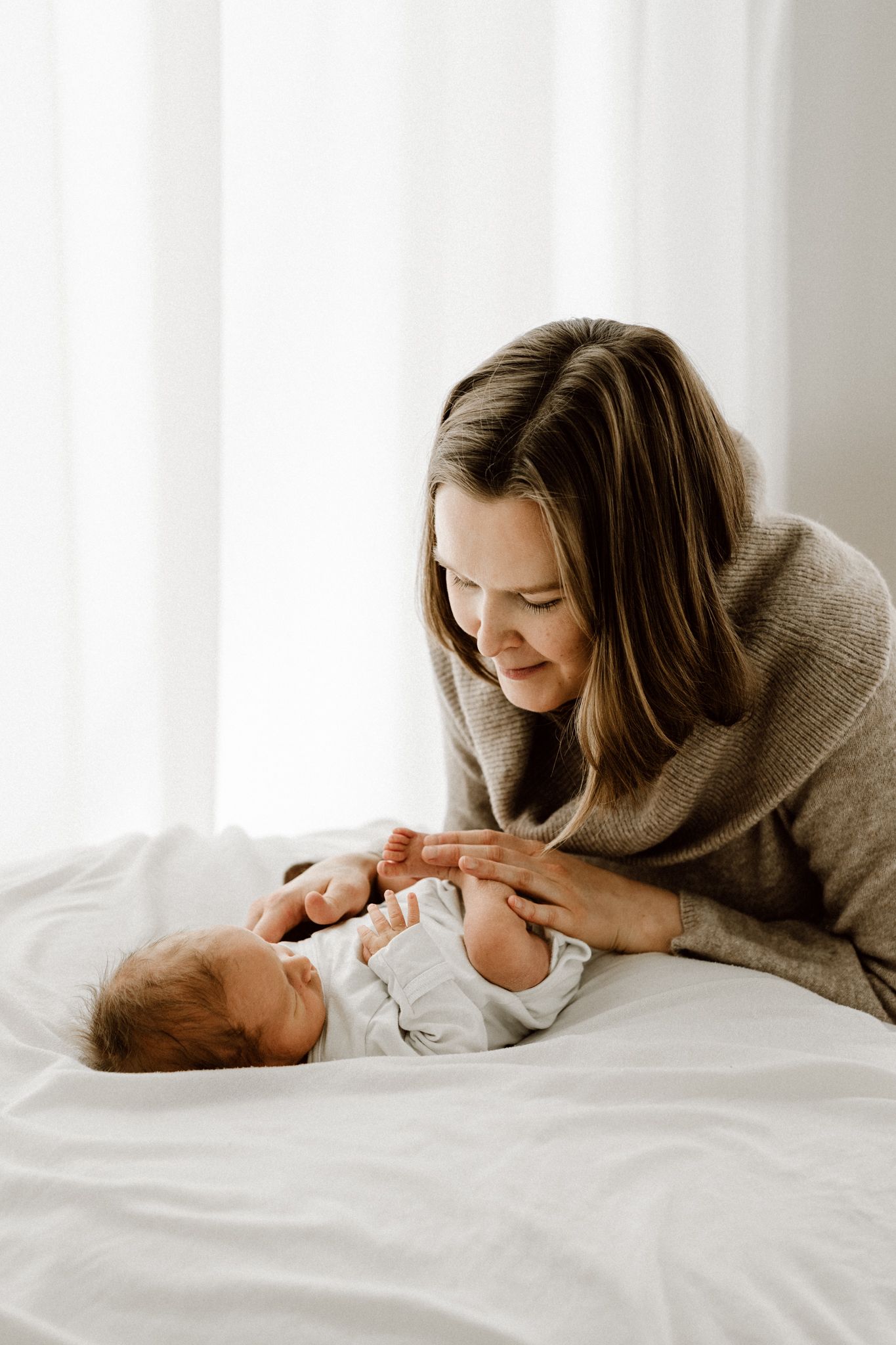 Smiling mother in brown sweater leaning over her newborn baby lying on a white bed.