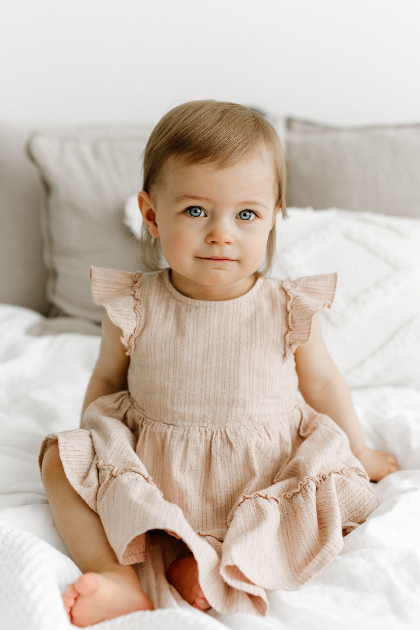 Sweet blue-eyed baby girl in ruffled beige dress sitting on white bed looking at camera.