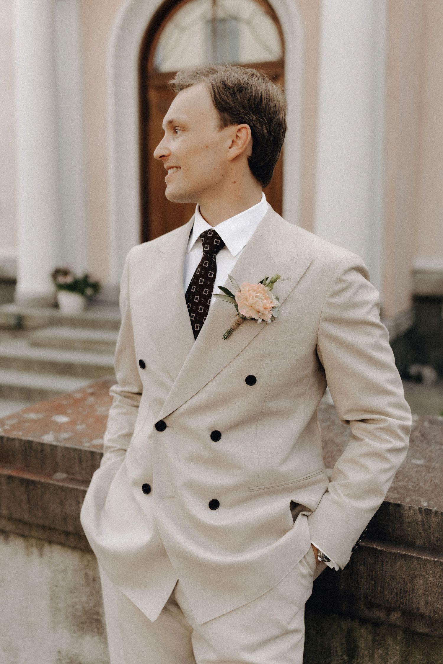 Dapper groom in a beige double-breasted suit with a floral boutonniere smiling outdoors near a classical building.