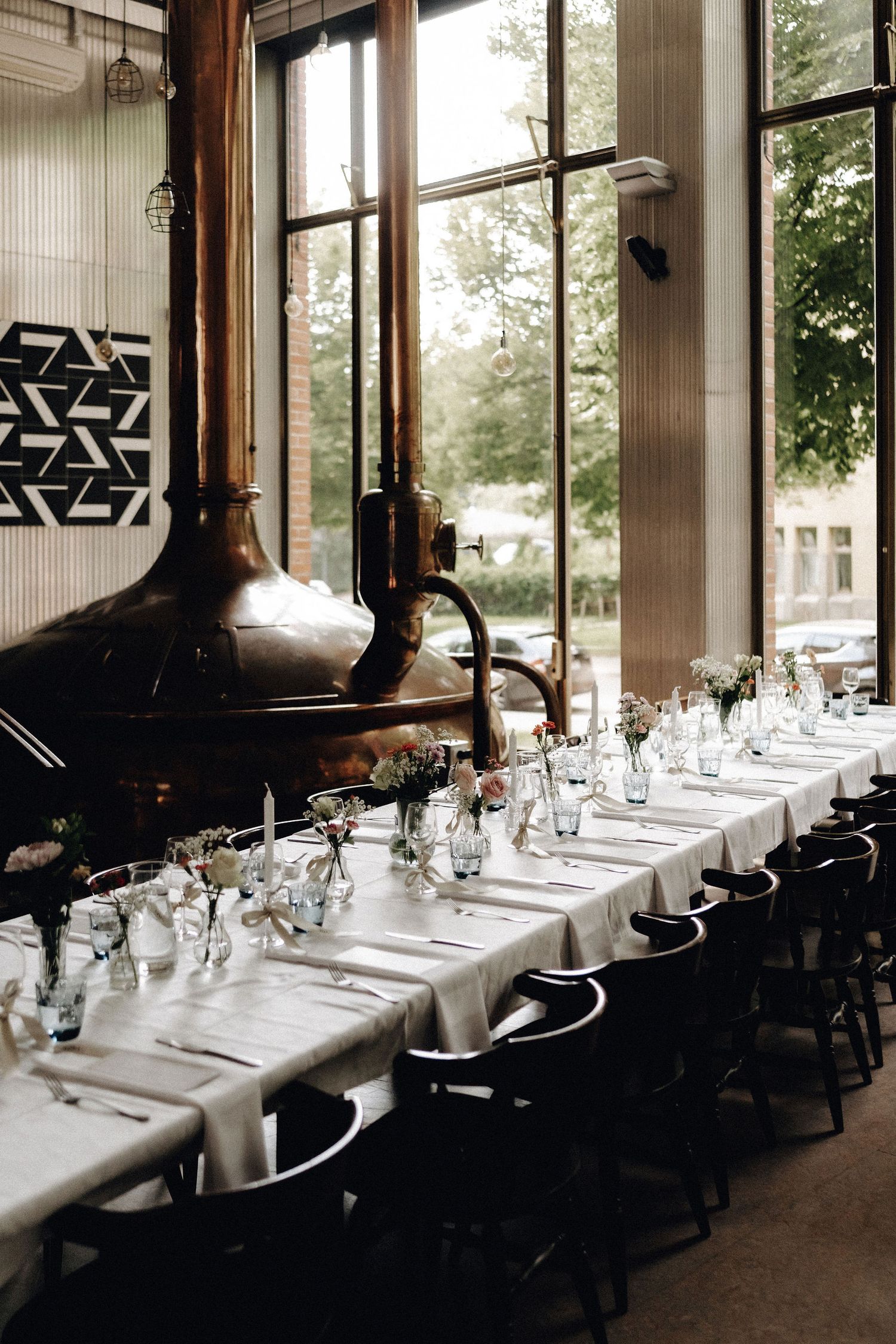 Long wedding reception table set with white linens and flowers beside large copper brewery tanks.