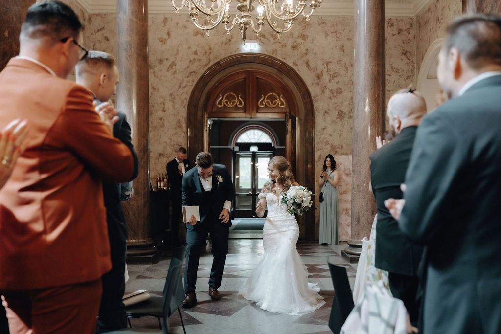 Bride and groom make grand entrance into ornate marble reception hall with chandelier and guests.