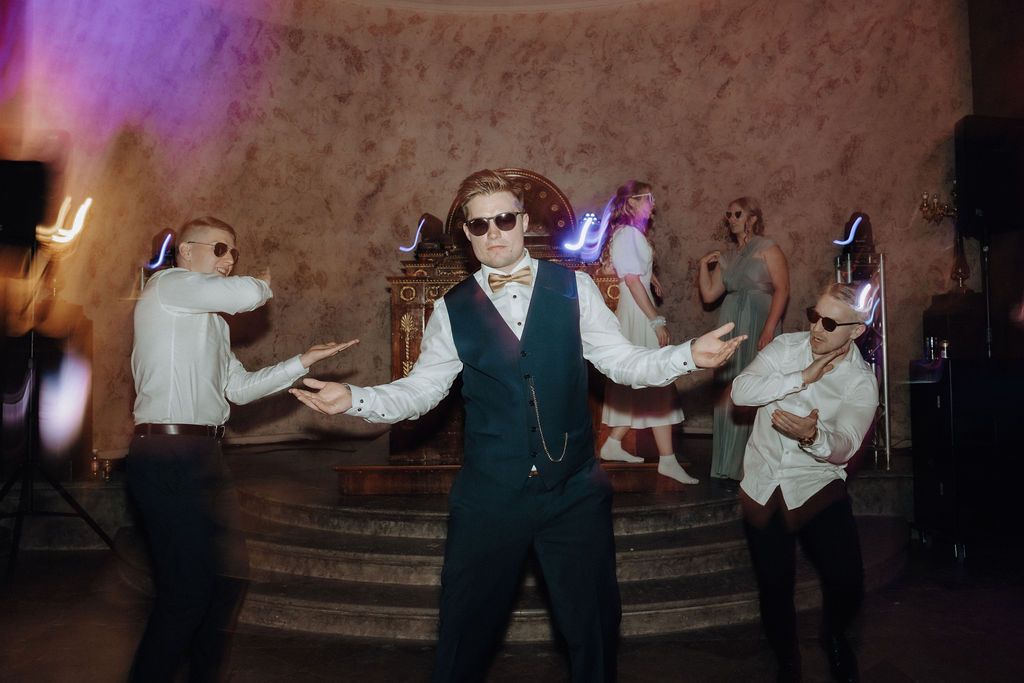 Groom and groomsmen wearing sunglasses perform energetic choreographed dance with colorful light effects.