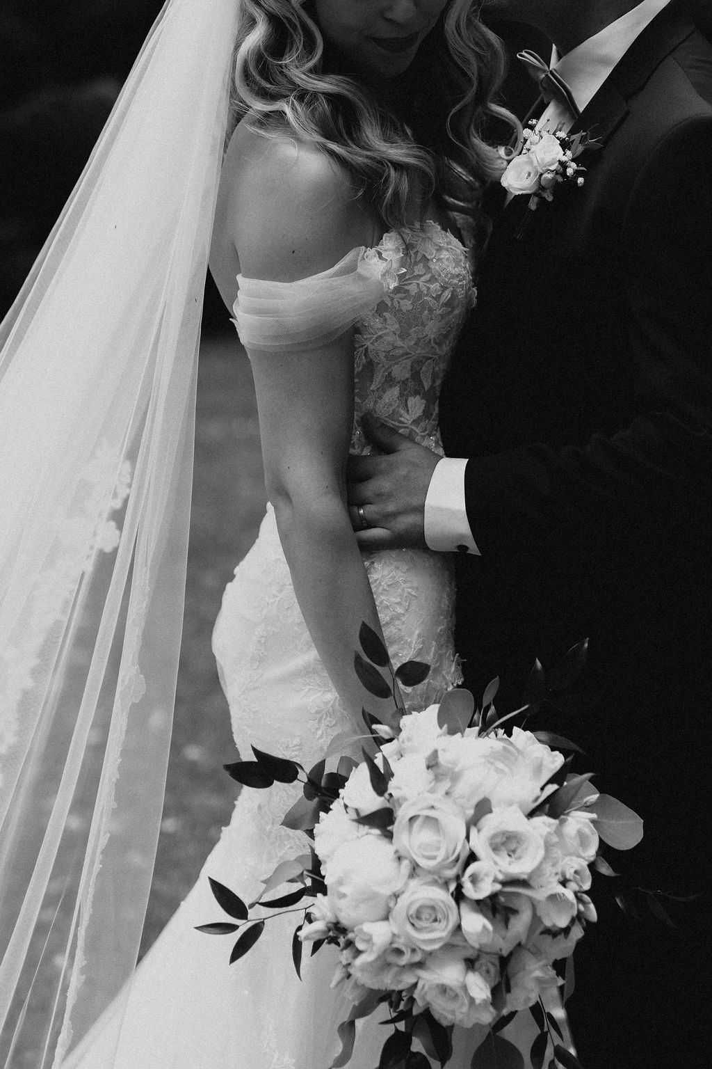 Black and white close-up of bride in lace off-shoulder gown holding white rose bouquet with groom embracing her.