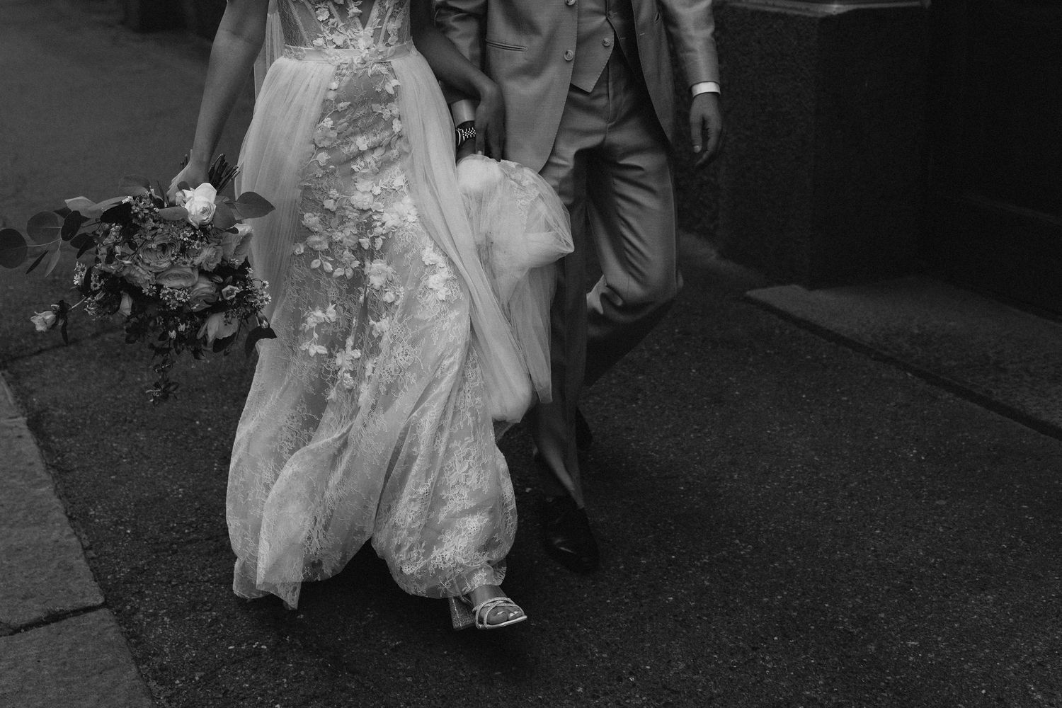 Black and white close-up of bride in lace gown and groom walking together on a sidewalk.