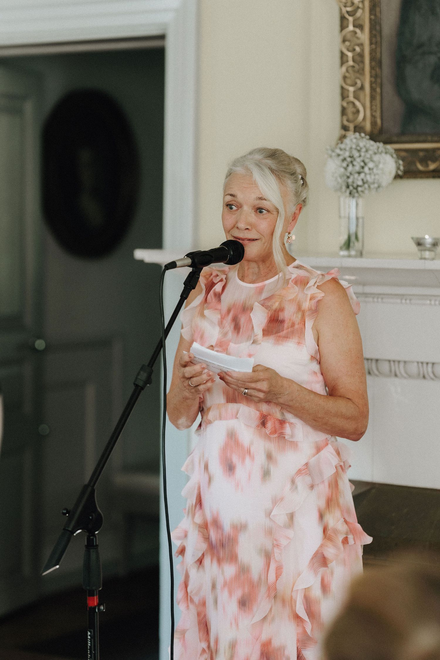 Elegant older woman in a floral dress delivering a heartfelt speech at a microphone during a wedding reception.