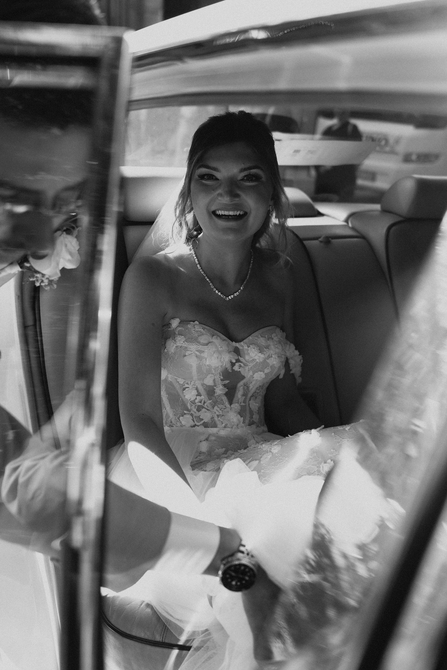 Black and white portrait of a laughing bride seated inside a classic white wedding car.