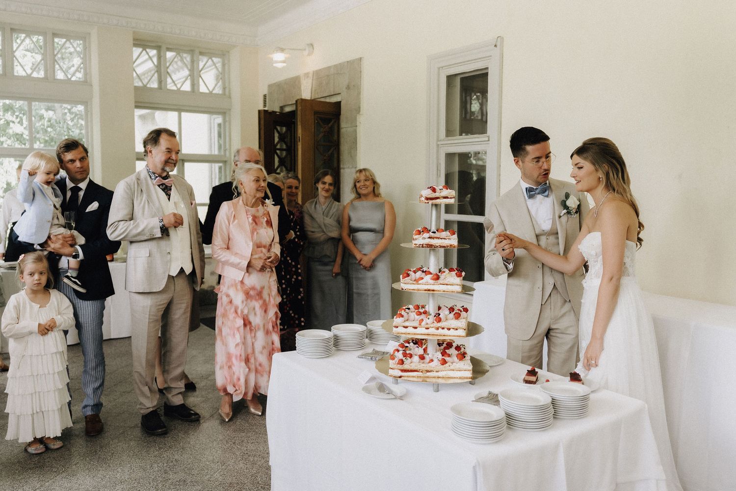 Wedding couple cutting a tiered strawberry cake surrounded by smiling family guests in a bright venue.