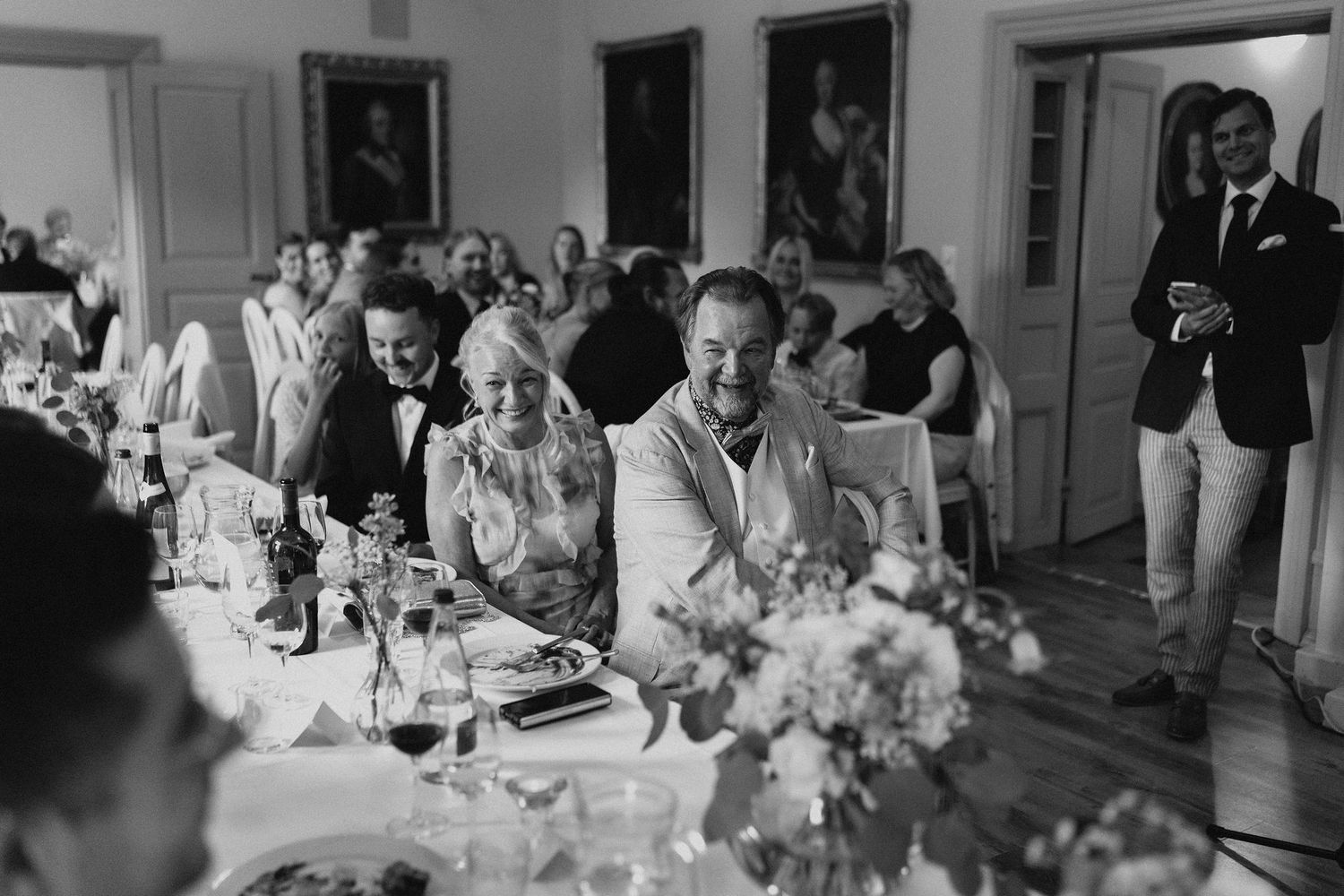Black and white photo of wedding guests laughing and enjoying speeches at a long reception table.