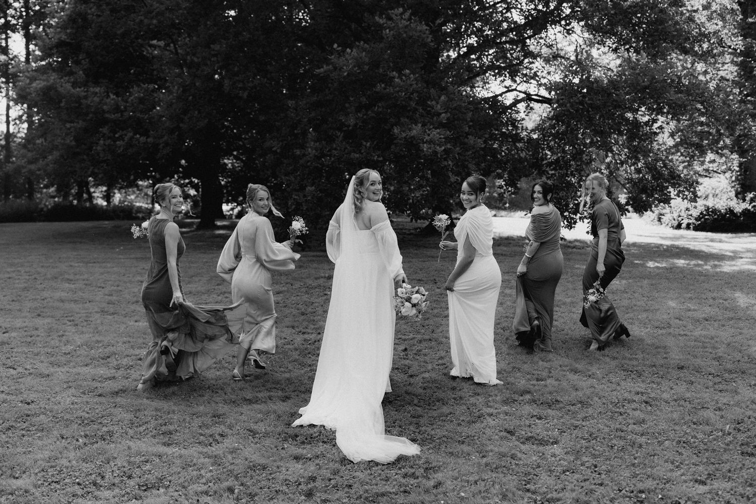 Bride in white gown walks with bridesmaids across green lawn in elegant black and white wedding photo.