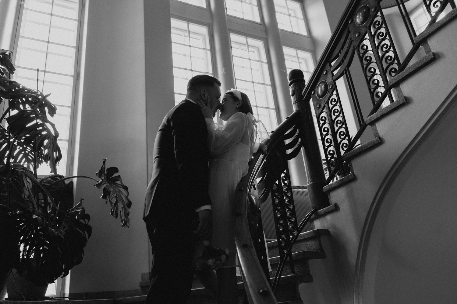 Mustavalkoinen valokuva pariskunnasta, joka suutelee romanttista suudelmaa elegantissa portaikossa, jossa on koristeelliset rautakaiteet.