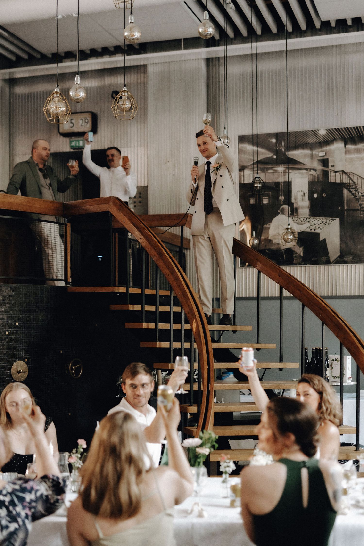 Groom in beige suit delivering speech on staircase while guests raise glasses in toast.