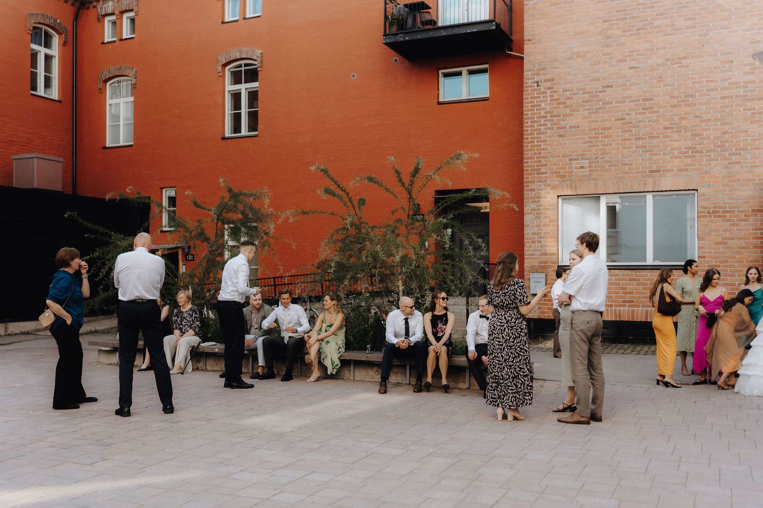 Wedding guests socialize in a sunny courtyard surrounded by red brick buildings.
