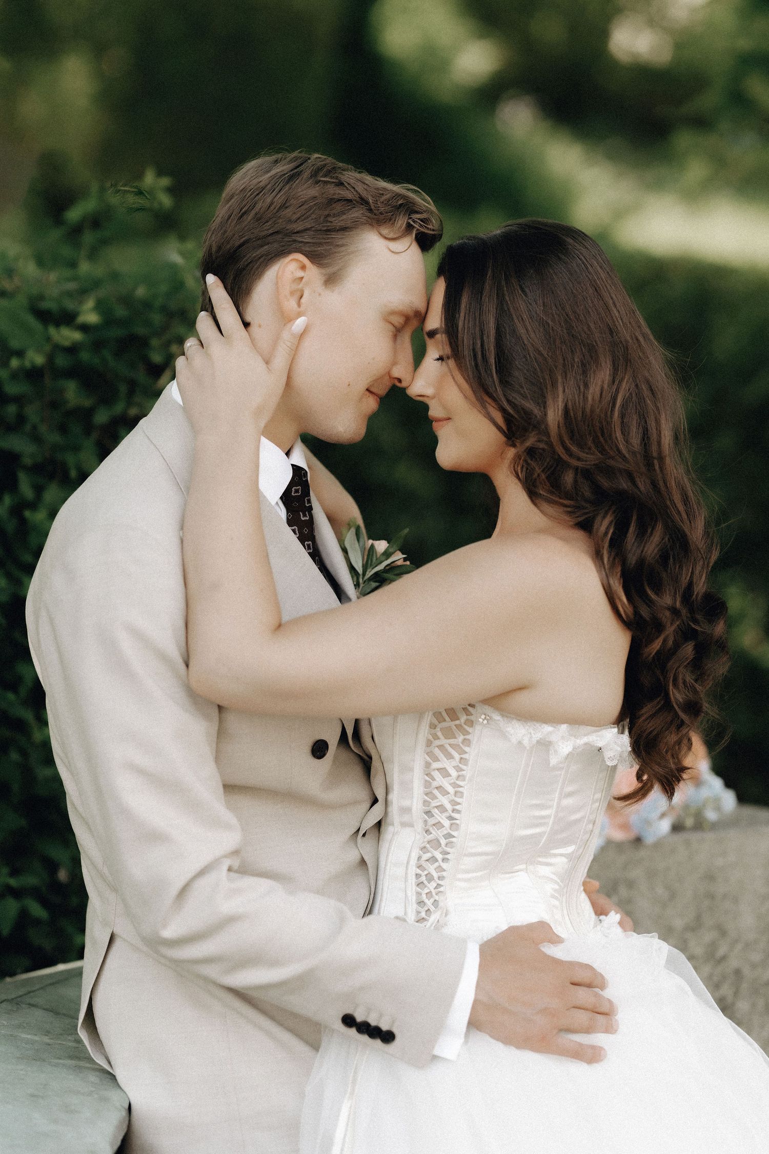 Bride and groom lean in for a kiss while embracing tenderly in a lush green outdoor garden setting.