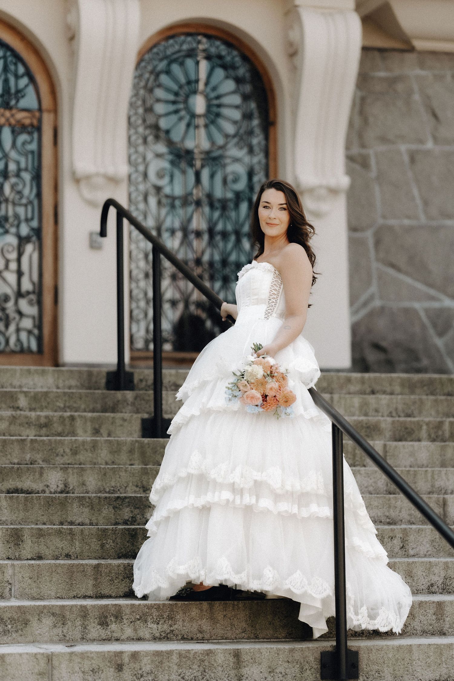 Elegant bride in layered white gown holding bouquet on stone steps before ornate iron door.