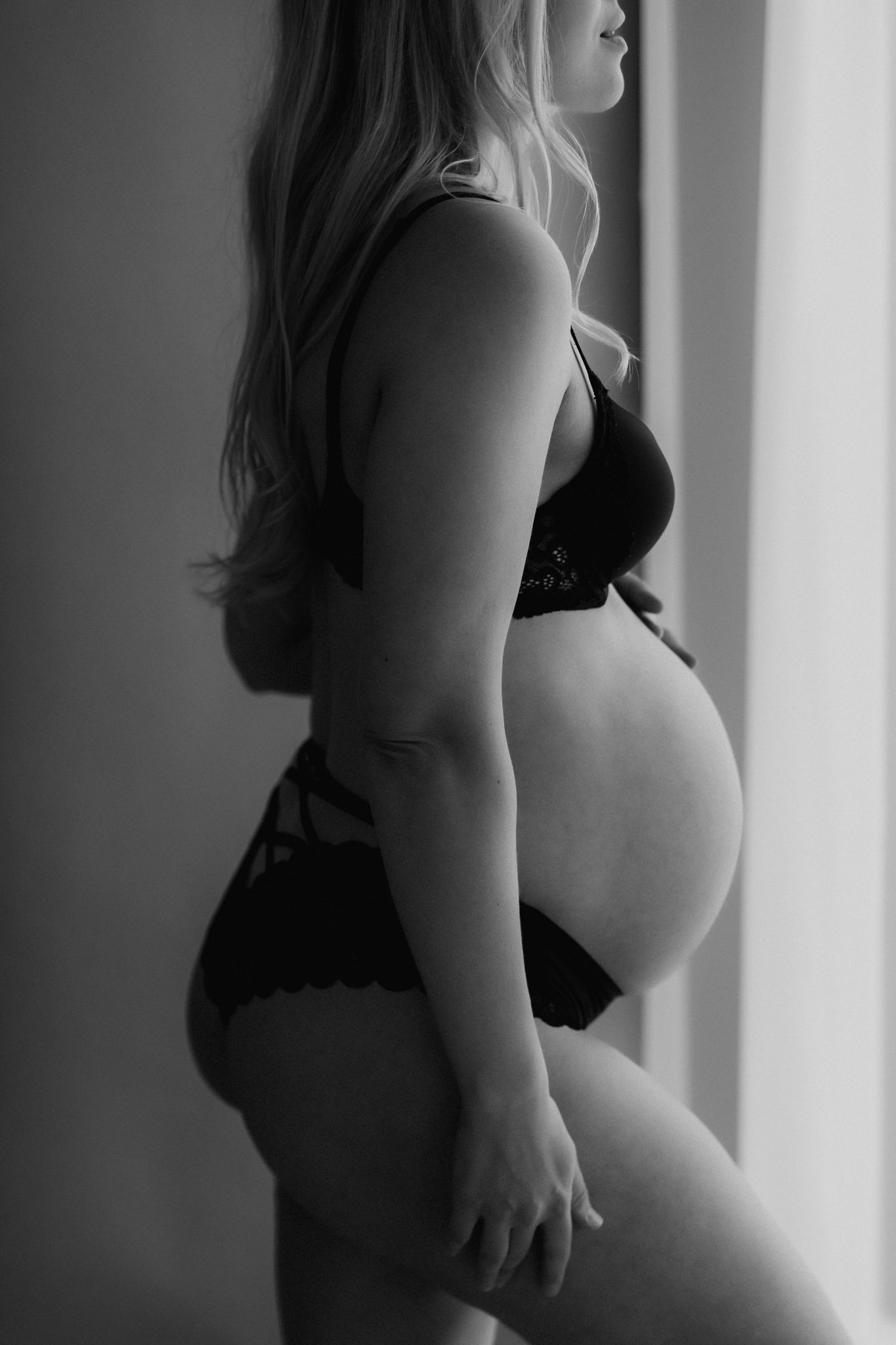 Pregnant woman in black lingerie showing her baby bump in profile near window, black and white maternity photo.