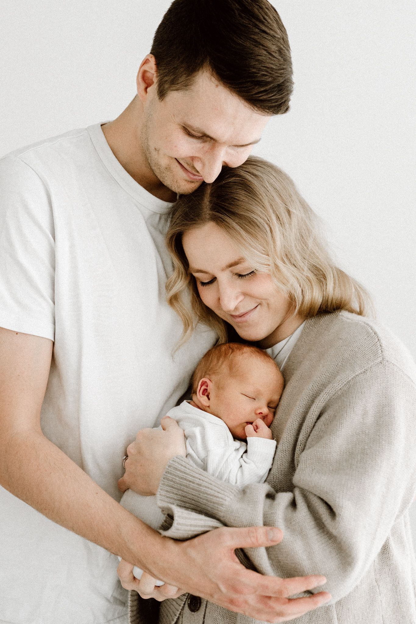 New parents in neutral tones tenderly embracing their sleeping newborn baby together.