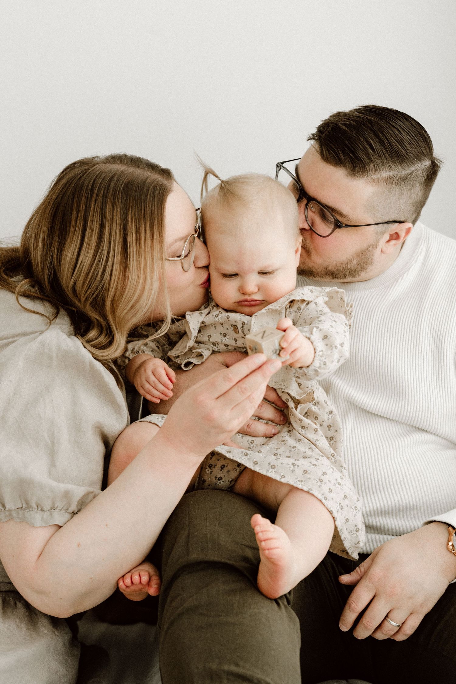 Two parents wearing glasses lovingly kiss their baby girl on the cheeks simultaneously in a bright studio.