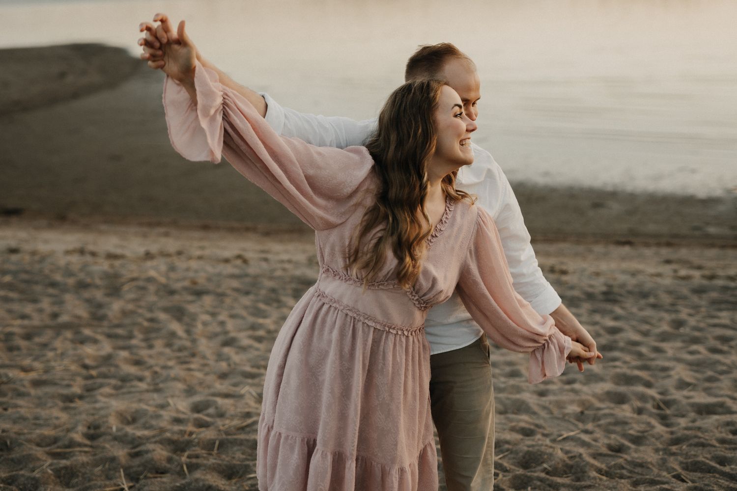 Couple dancing joyfully on sandy beach at sunset, woman in flowing pink dress laughing.