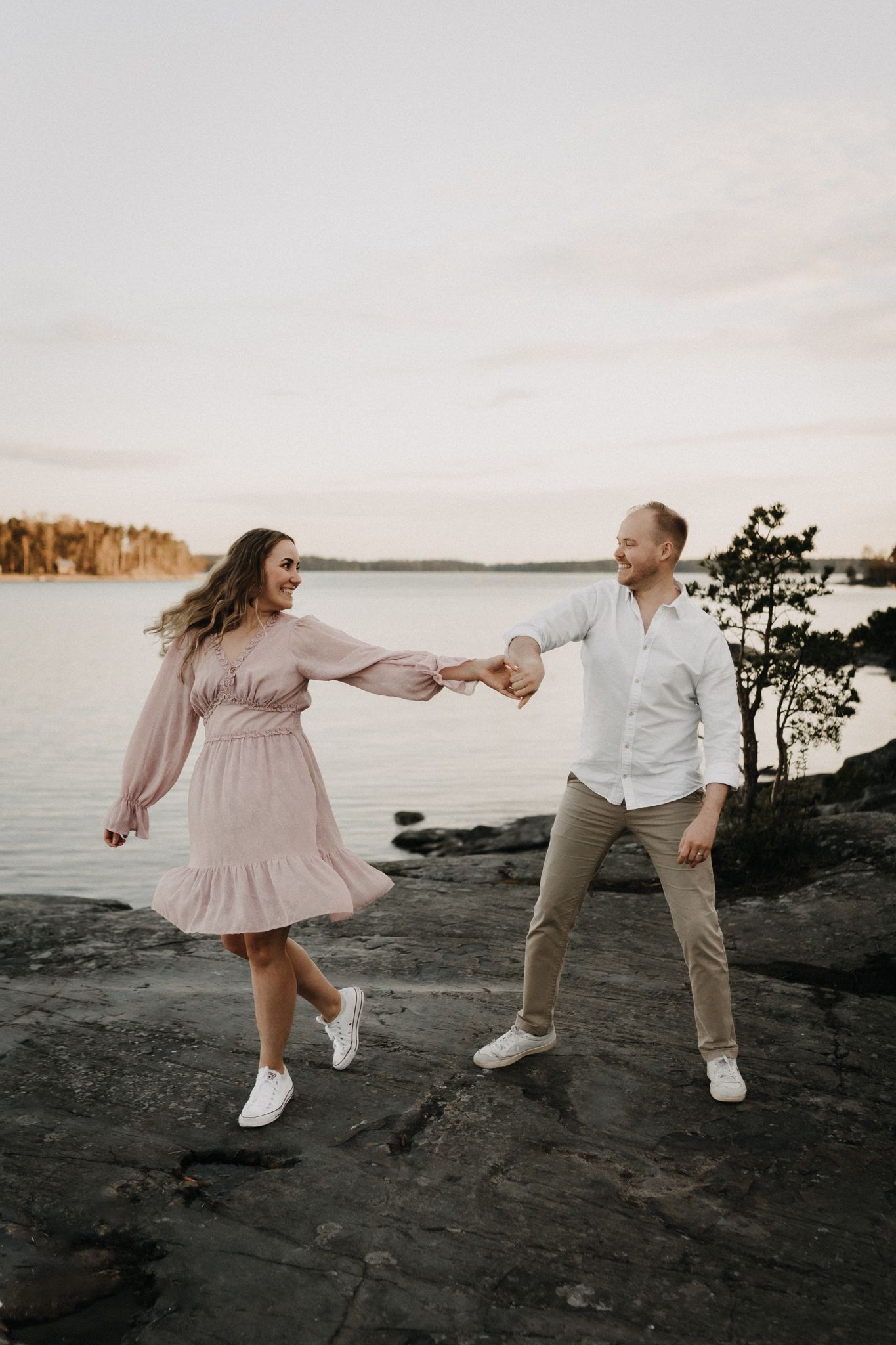 A joyful couple holds hands and dances on rocky shoreline at sunset, woman in pink dress and man in white shirt.