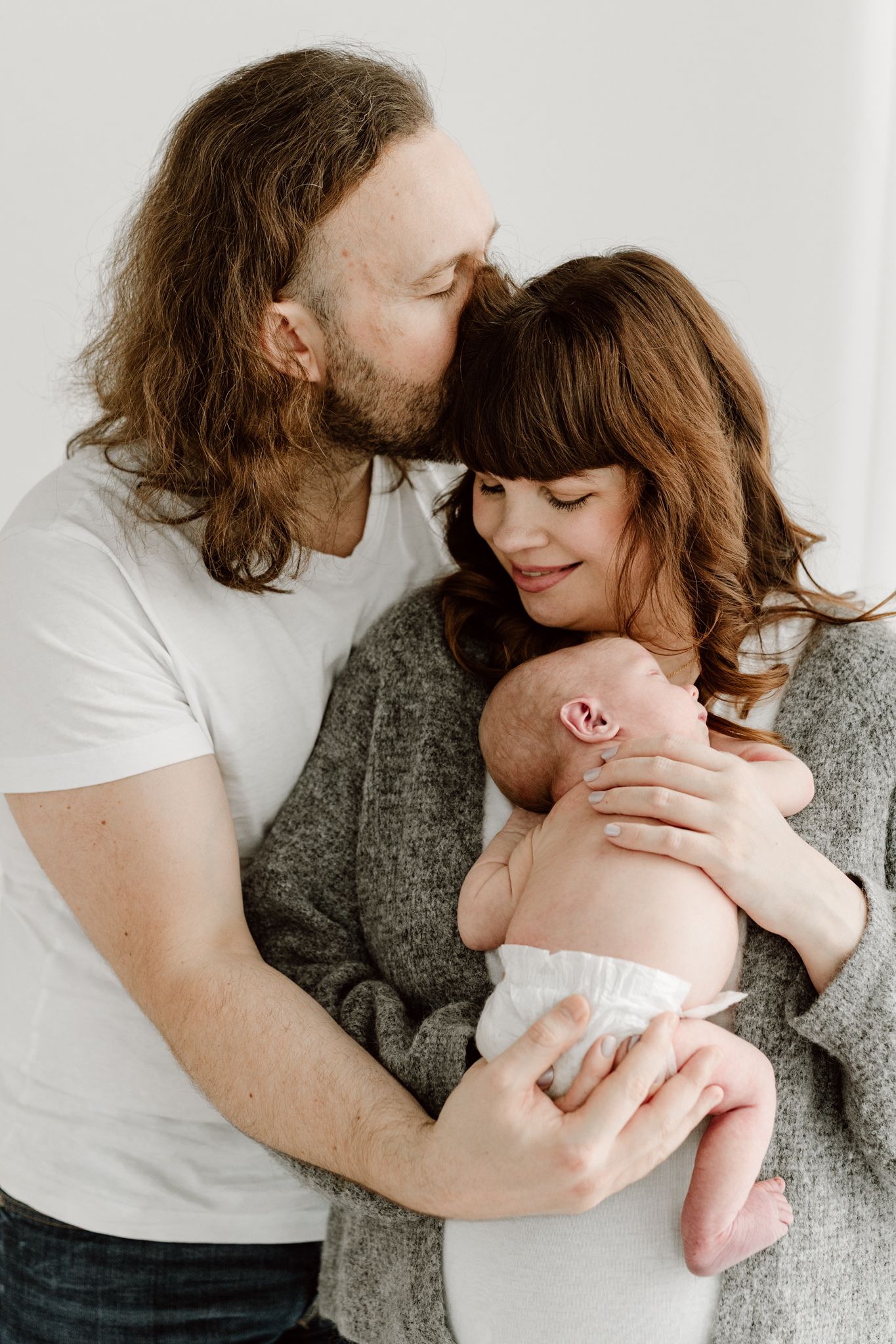 Father kissing mother on the cheek while she cradles their newborn baby in a bright studio.