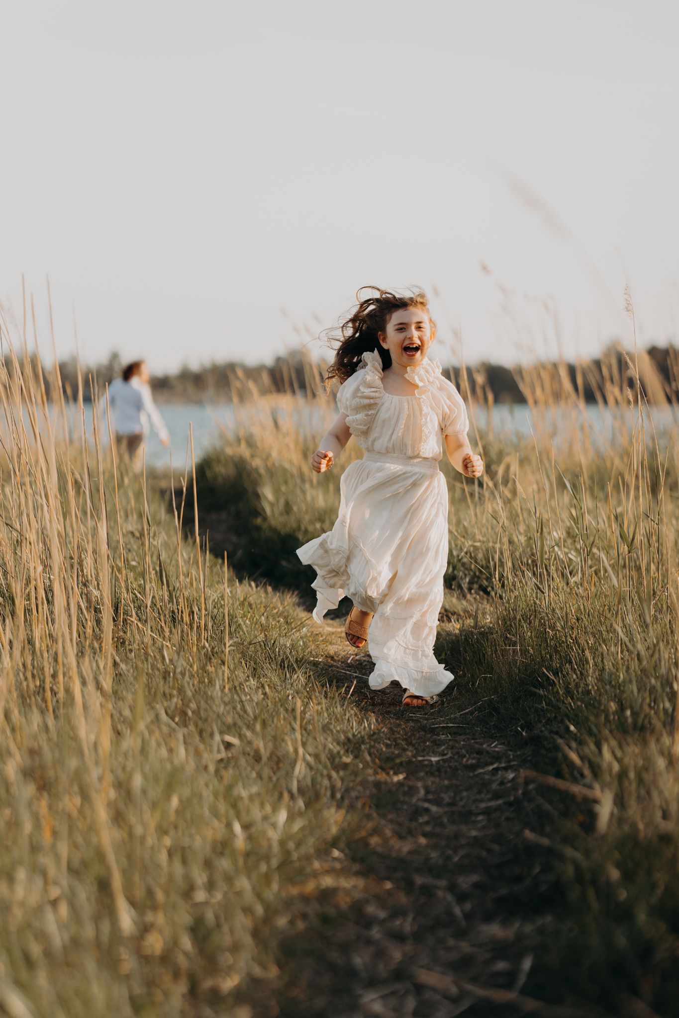 A joyful girl in a white dress runs laughing along a grassy path near a lake at golden hour.