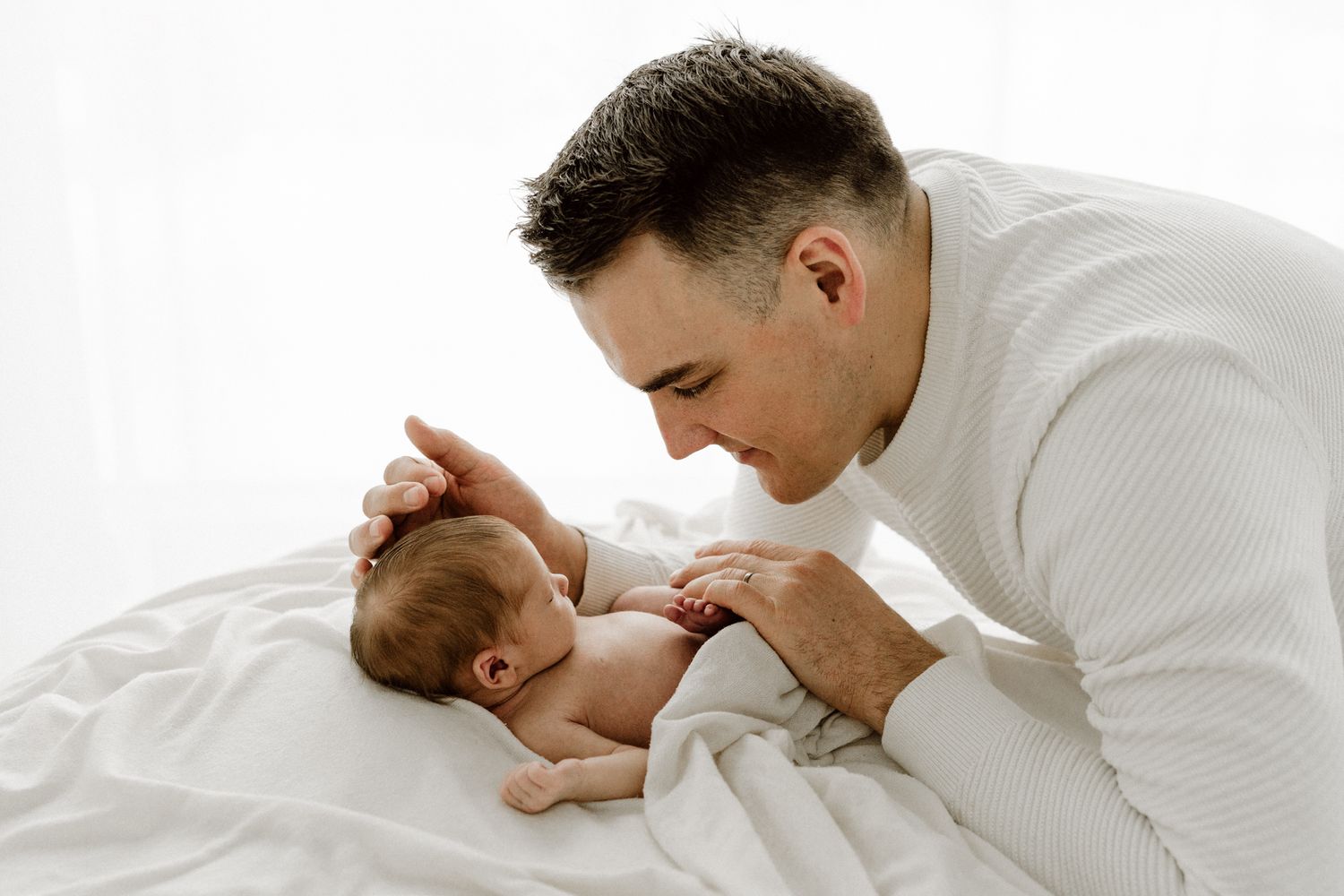 Devoted father in white sweater leans over to gently interact with his newborn baby on a white blanket.