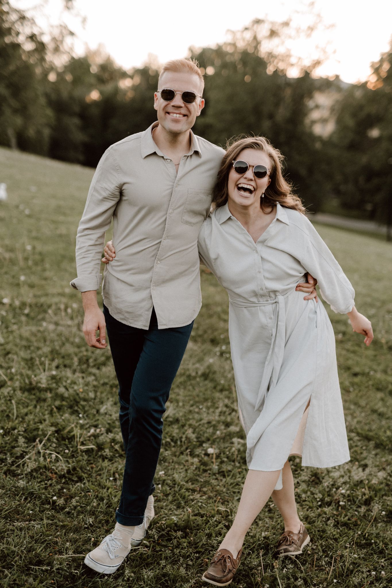 Joyful couple in sunglasses walking arm in arm on a grassy hill at sunset.