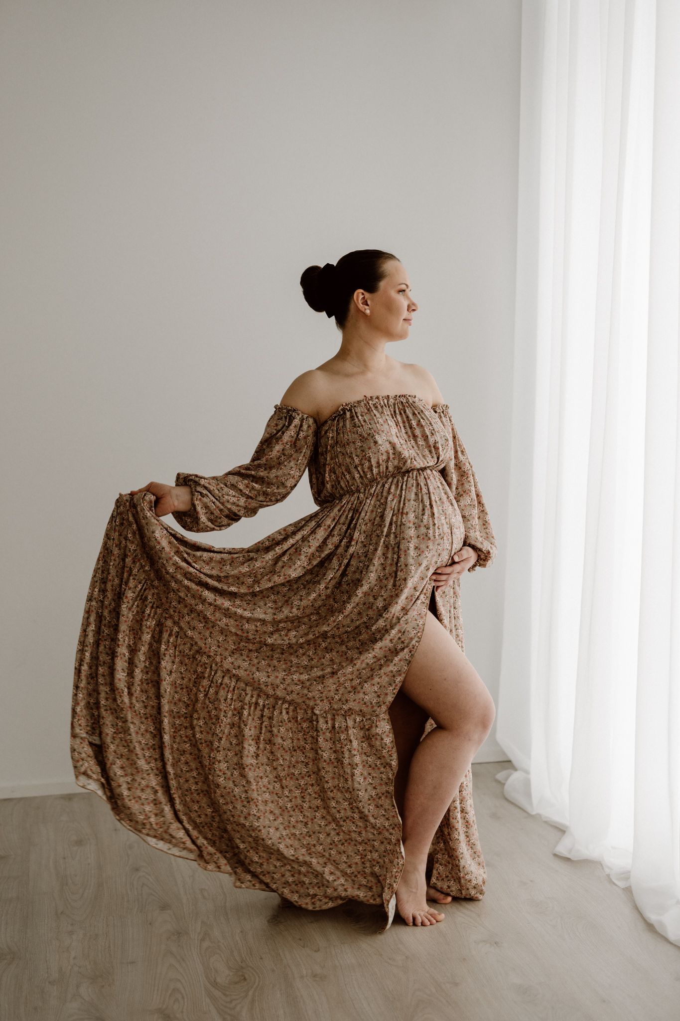 Pregnant woman in flowing floral off-shoulder maxi dress twirling near white curtains barefoot.