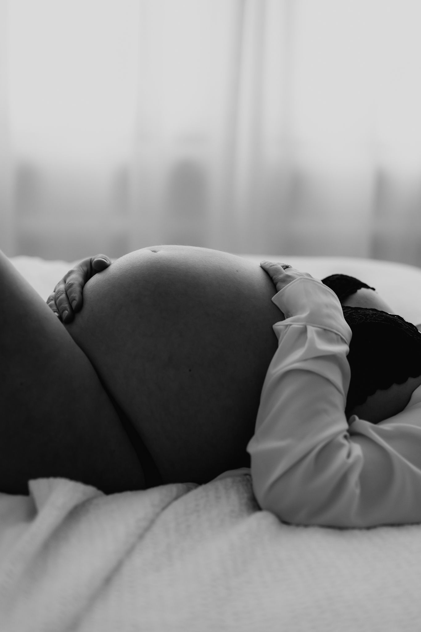 Black and white photo of pregnant woman lying on bed cradling her bare baby bump with both hands.