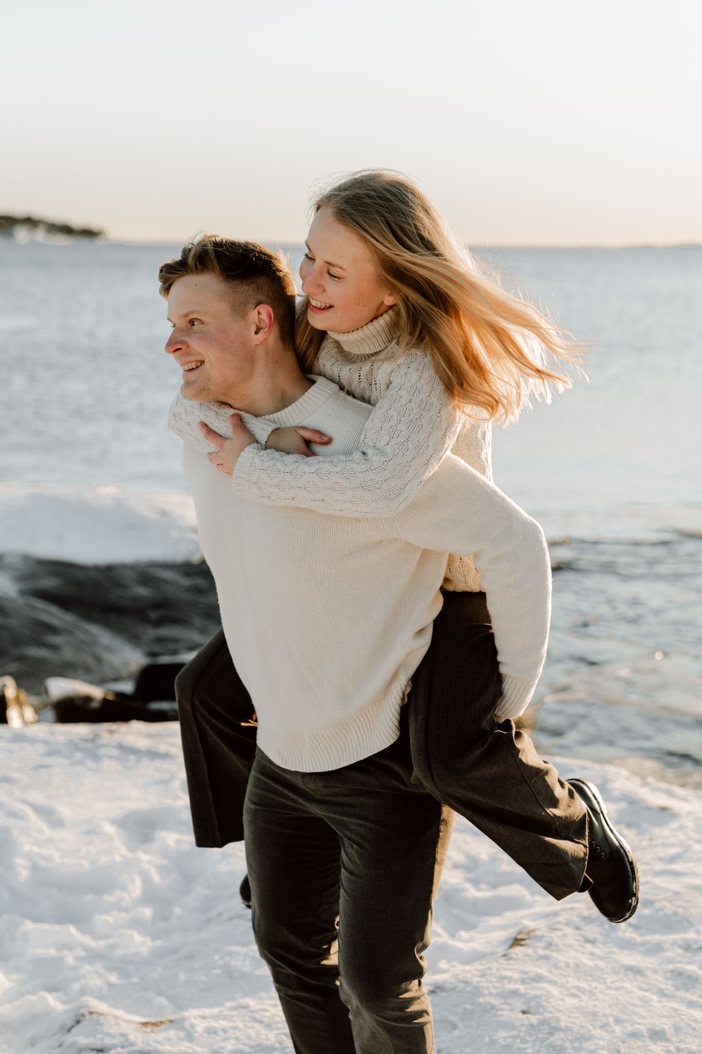 Laughing couple enjoy a piggyback ride together on a snowy coastal beach during winter sunset.
