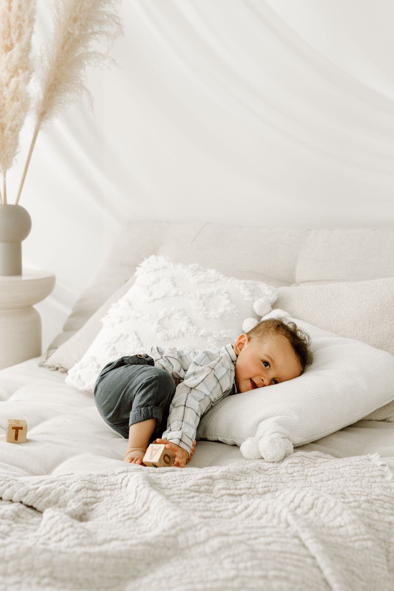 Baby boy in plaid shirt resting on white textured pillow on a boho-styled bed.