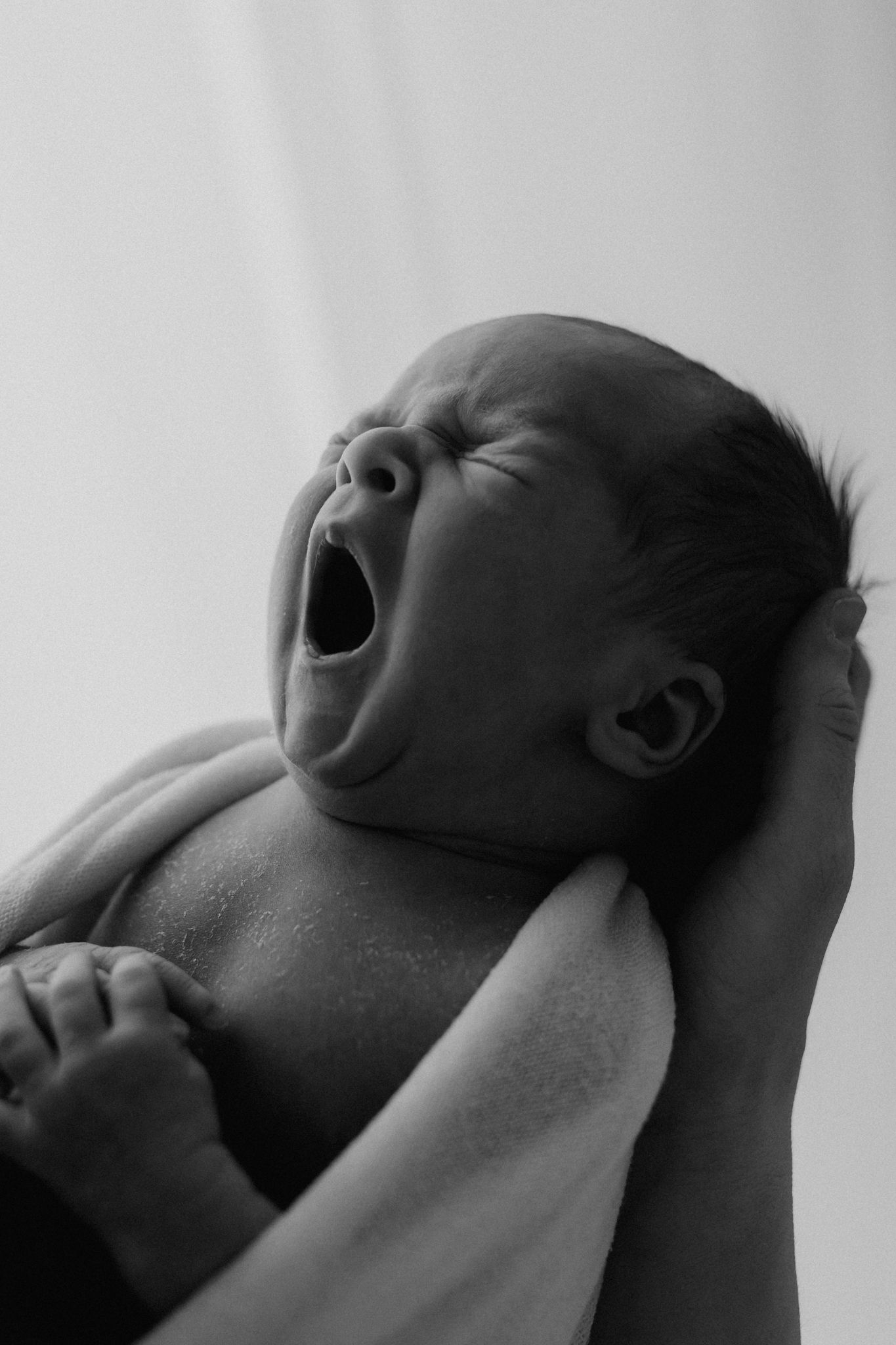 Black and white photo of a newborn baby yawning while being held by adult hands.