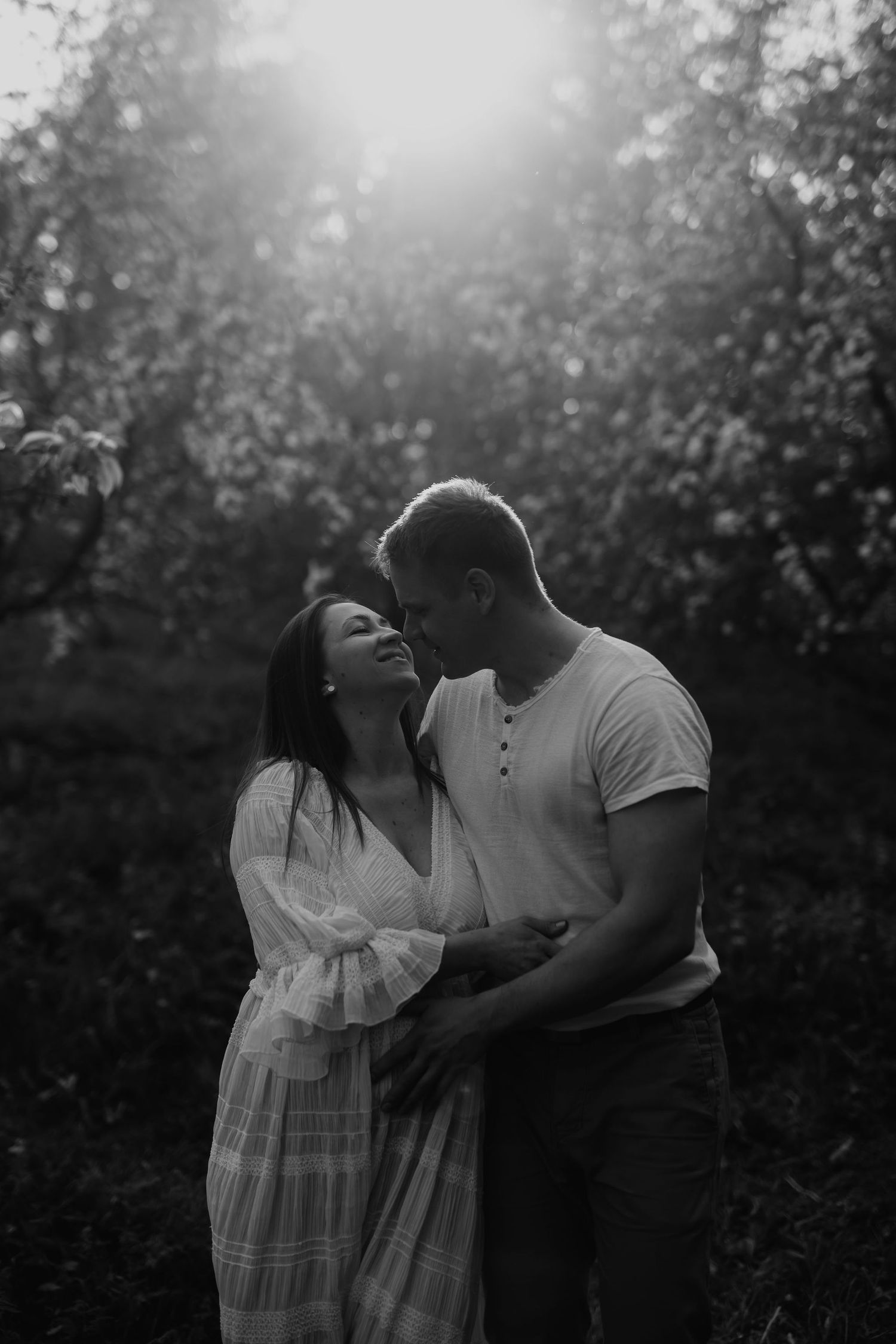 Black and white romantic photo of smiling couple embracing in sunlit blooming orchard at golden hour.