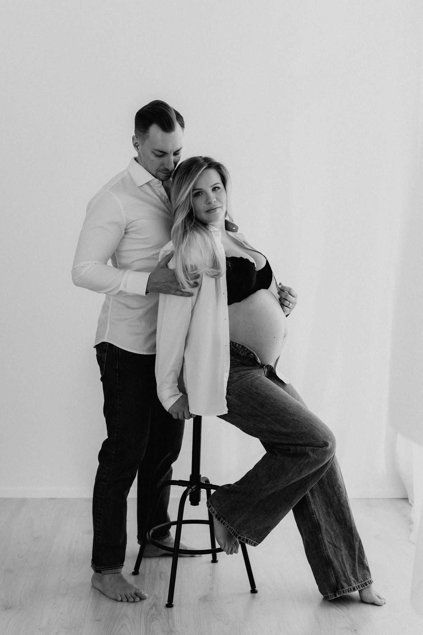 Black and white studio maternity photo of couple, pregnant woman seated on stool with partner.