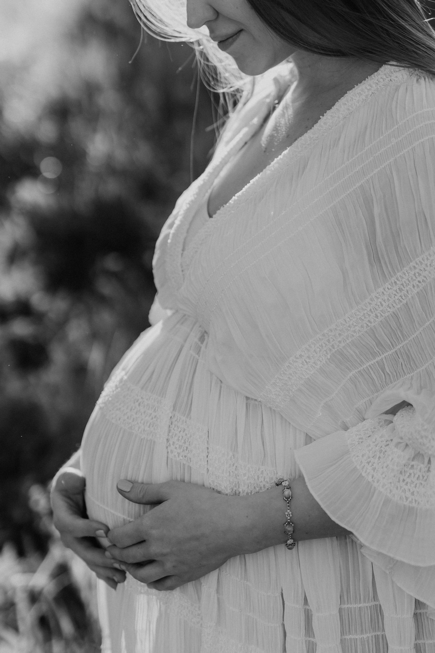 Black and white photo of pregnant woman in white dress cradling baby bump outdoors.