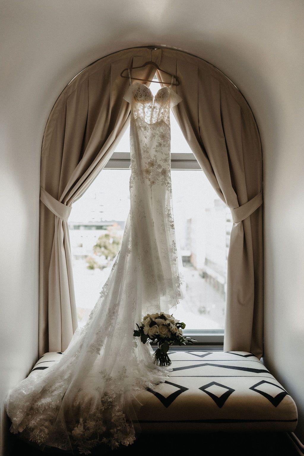 Elegant lace wedding dress hanging in arched window with bridal bouquet on cushioned seat below.