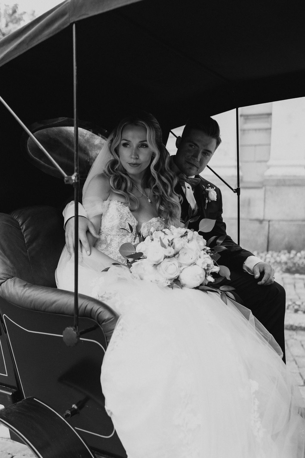 Black and white photo of bride and groom sitting together in a vintage carriage on wedding day.