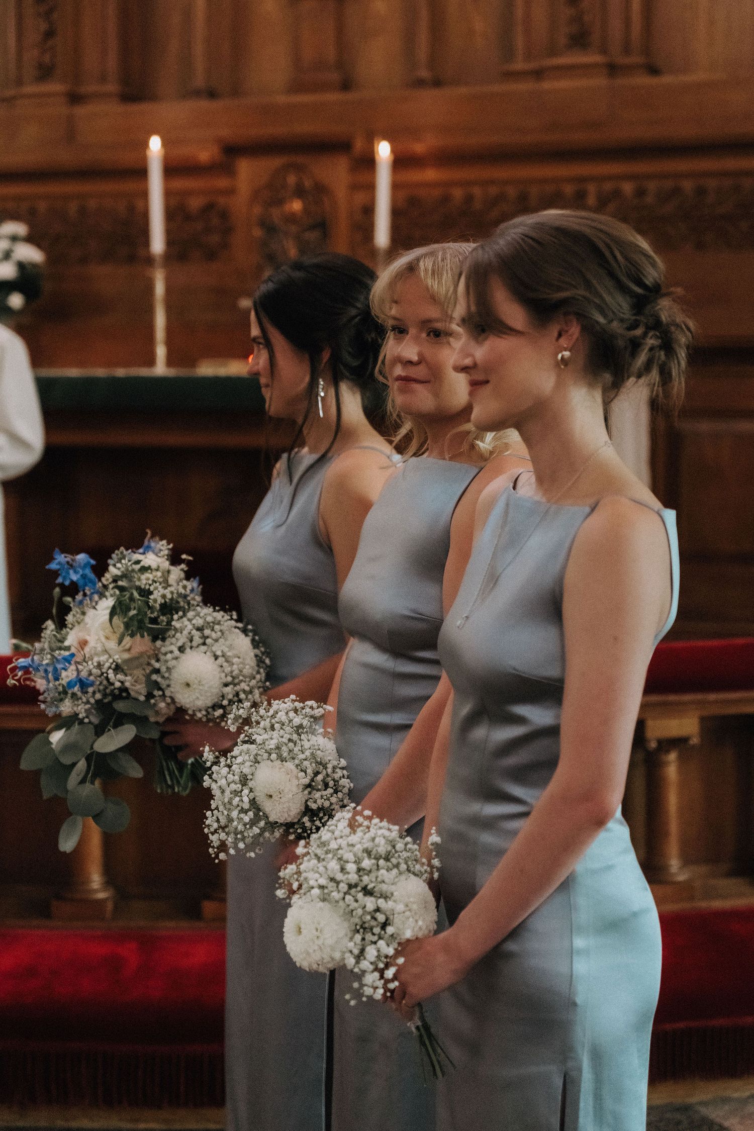 Three bridesmaids in matching silver-blue gowns holding white floral bouquets during church ceremony.