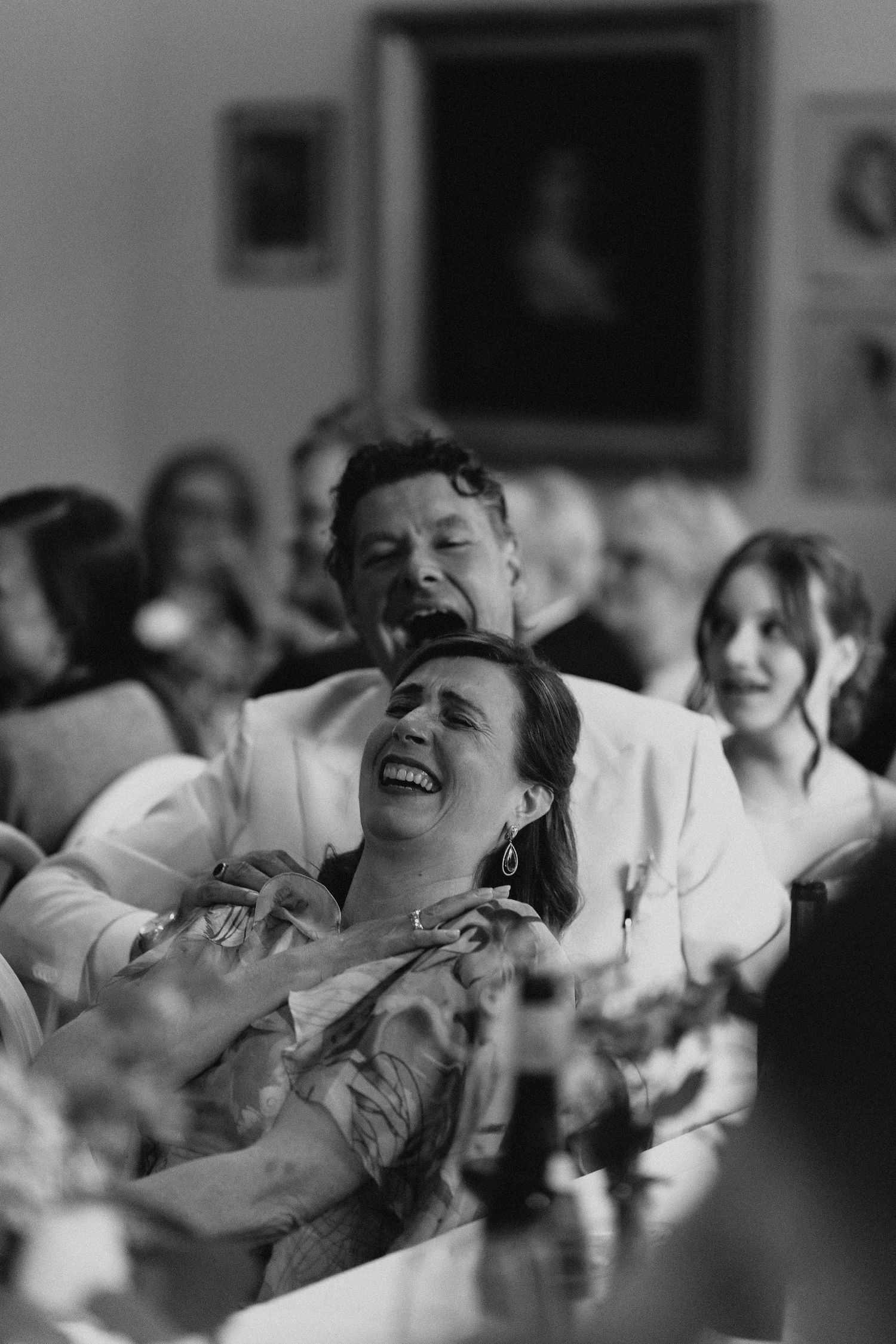 Joyful wedding guests laughing and embracing at dinner table during reception speeches, black and white.