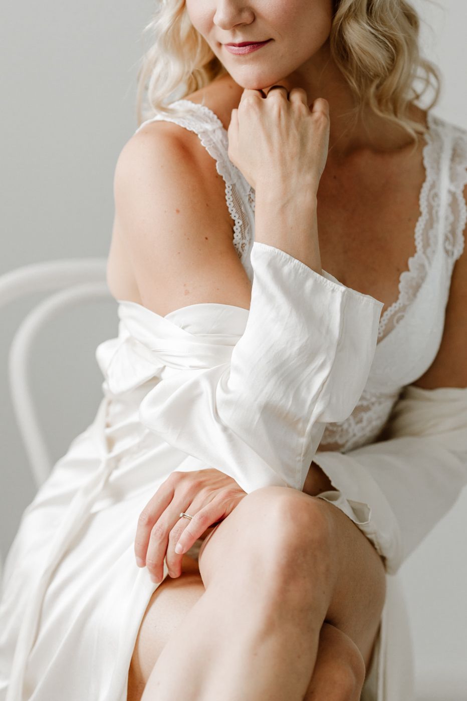 Woman in white lace lingerie and robe sitting on chair smiling softly.