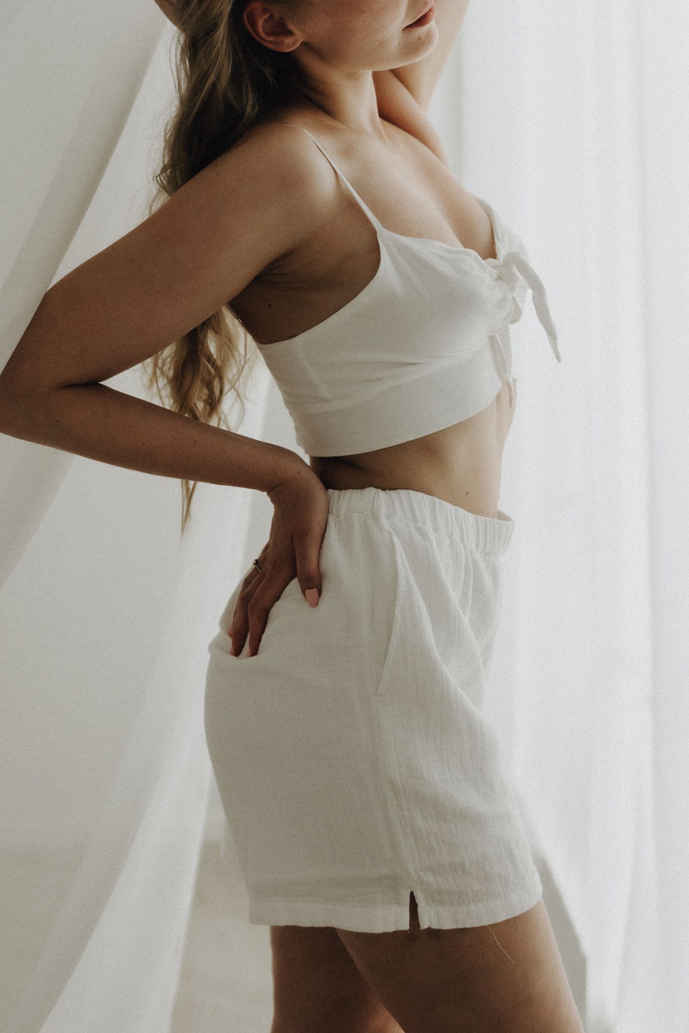 Woman in white linen crop top and matching shorts posing near sheer curtains.