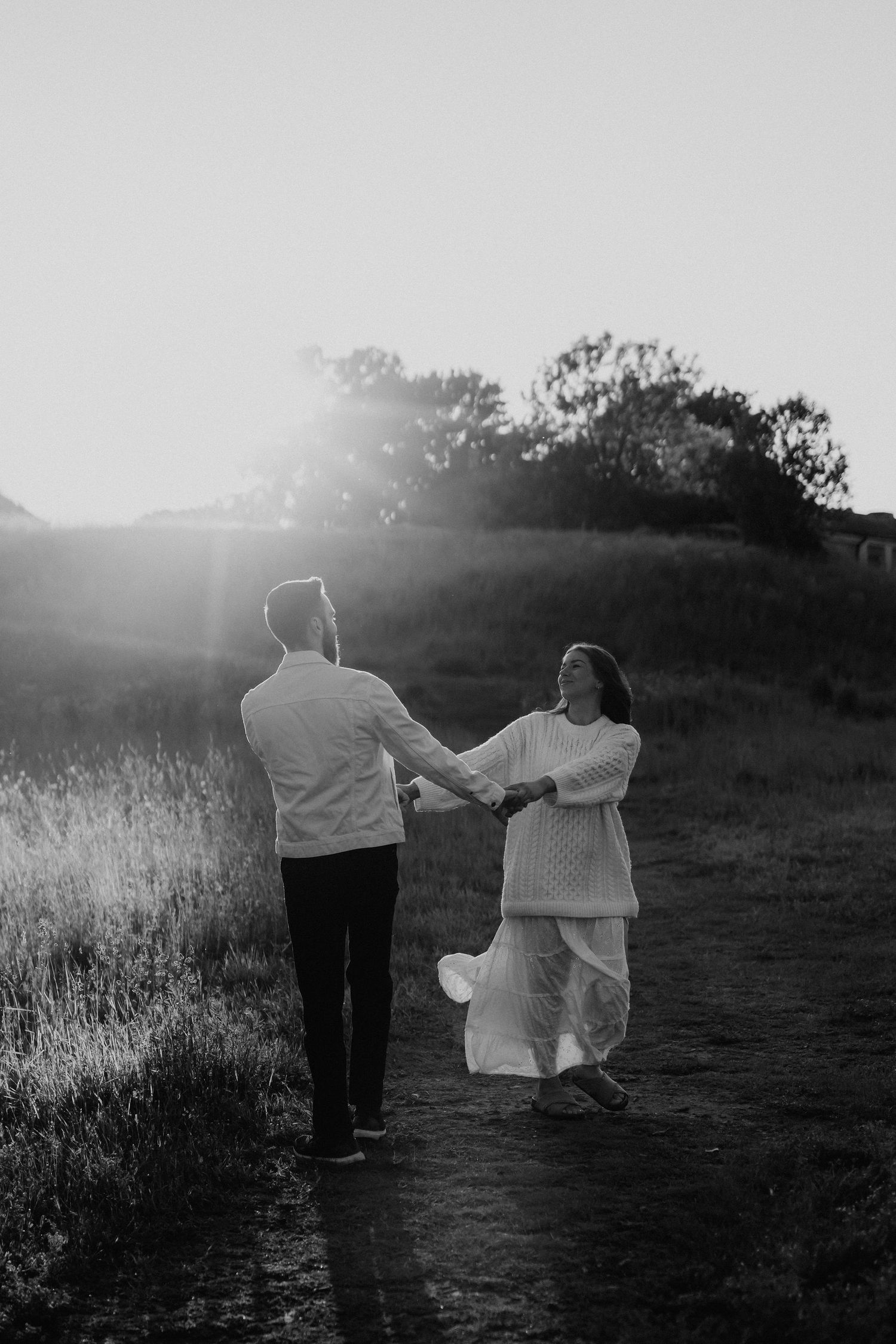 Black and white photo of a couple holding hands walking along a path at golden hour.
